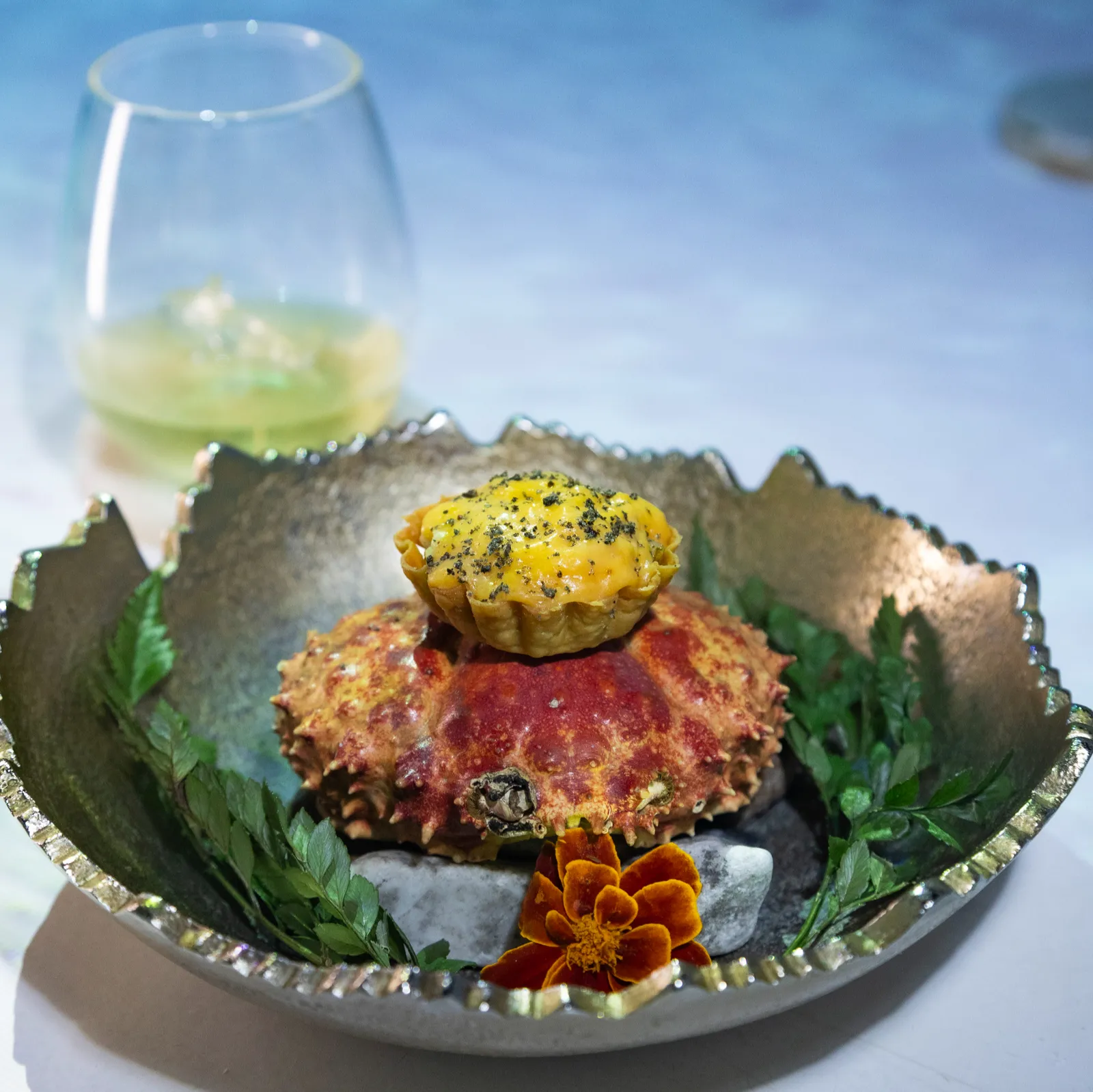 Plato de Frenessí: un cangrejo gratinado con una tartaleta amarilla encima, sobre piedras, hojas verdes y una flor naranja, junto a un vaso con bebida.