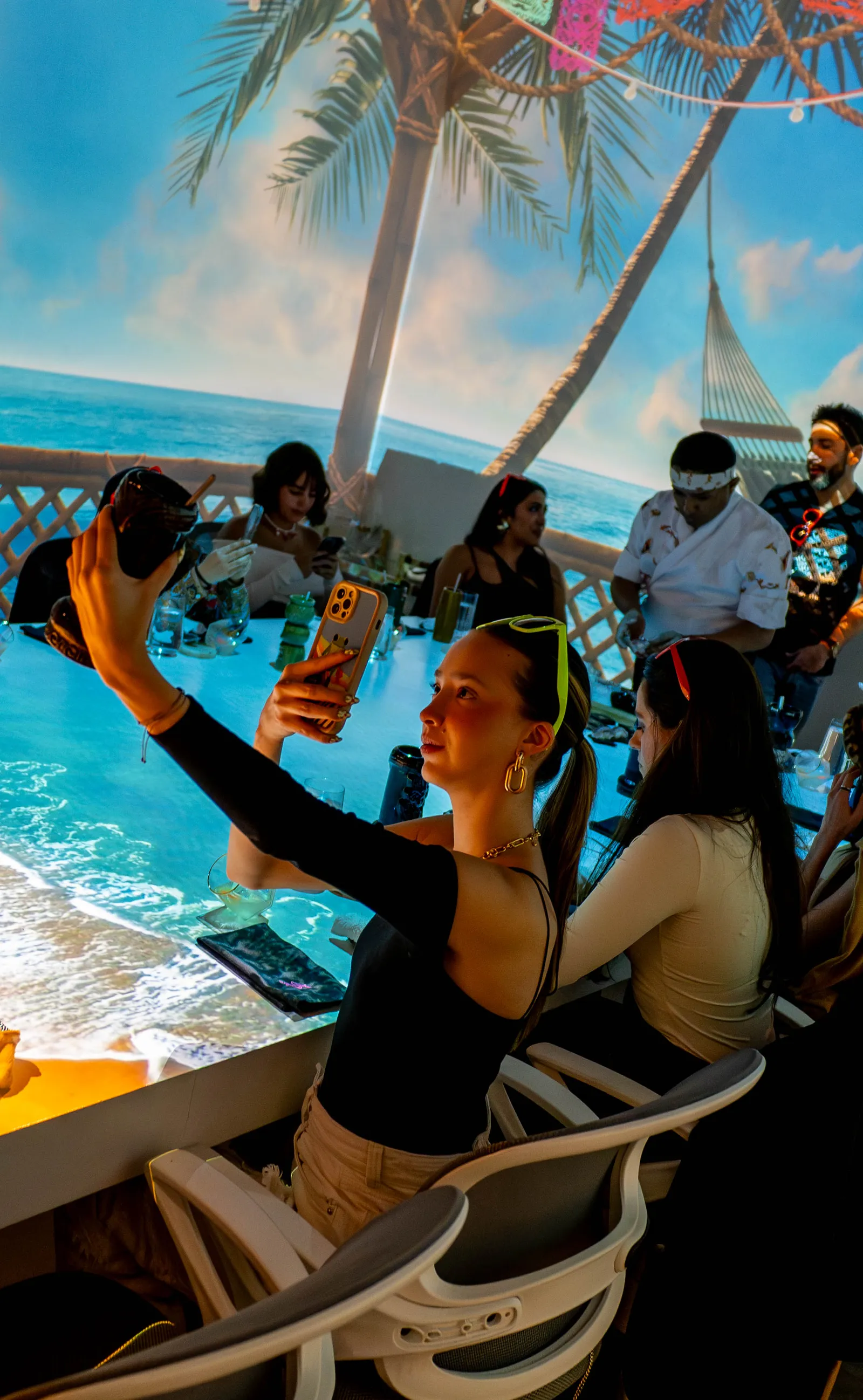 Una comensal se toma una selfi en la mesa, con una playa de palmeras proyectada detrás y los cocineros al fondo.